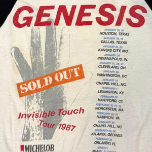 May include: Cream-colored vintage t-shirt featuring the band name "GENESIS" in red, with a hand graphic and tour dates for the "Invisible Touch Tour 1987." The shirt also has the "SOLD OUT" text.