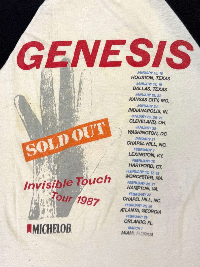 May include: Cream-colored vintage t-shirt featuring the band name "GENESIS" in red, with a hand graphic and tour dates for the "Invisible Touch Tour 1987." The shirt also has the "SOLD OUT" text.