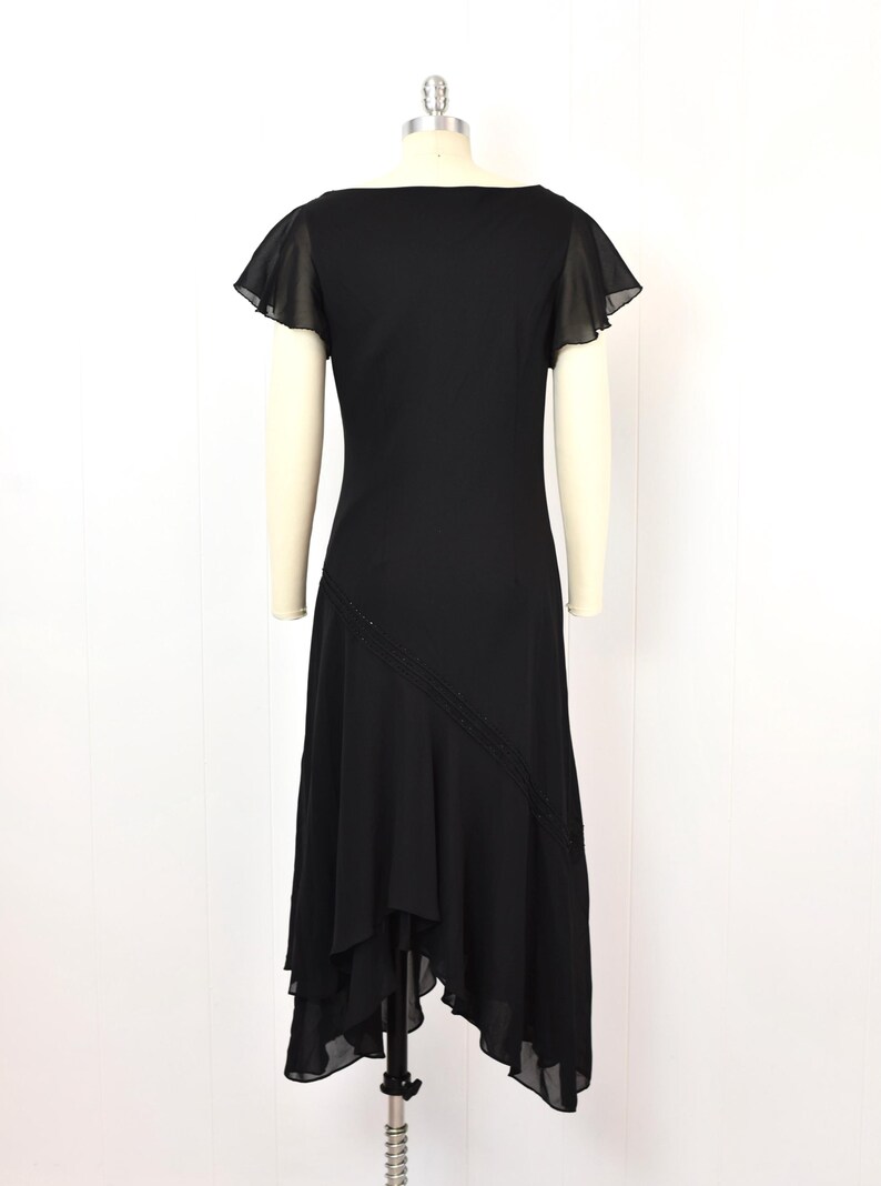 May include: A black dress with a square neckline and short, sheer flutter sleeves. The dress has an asymmetrical hemline and a diagonal beaded detail. The dress is displayed on a mannequin against a white background.