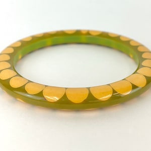 May include: Olive green bangle bracelet with a repeating pattern of light orange half-circle shapes. The bracelet is made of a glossy material and has a smooth, rounded surface. A stylish accessory.