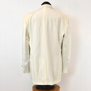 May include: A cream-colored blazer displayed on a mannequin. The back view shows the tailored fit, with a classic notched lapel and buttoned cuffs. The jacket is made of a smooth fabric, suggesting a formal or semi-formal style.