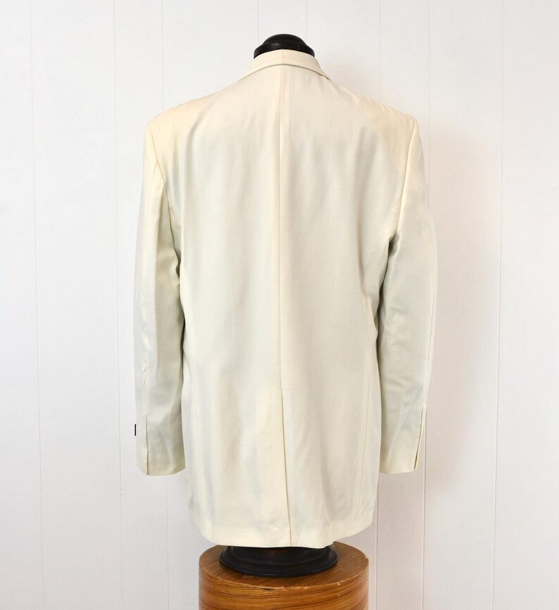 May include: A cream-colored blazer displayed on a mannequin. The back view shows the tailored fit, with a classic notched lapel and buttoned cuffs. The jacket is made of a smooth fabric, suggesting a formal or semi-formal style.