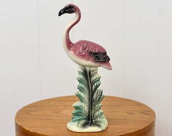 May include: A pink and white ceramic flamingo figurine standing on a green leafy base. The flamingo has a black beak and black eyes.
