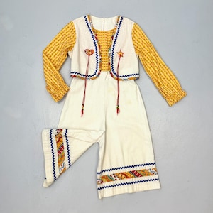 May include: A vintage children's outfit featuring a yellow floral print top with long sleeves and a white vest with blue trim. The outfit includes white pants with decorative trim. The vest has heart and star accents.