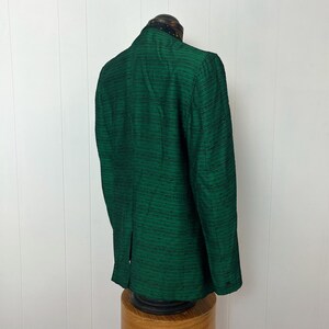 Może przedstawiać: Zielony, ciemny blazer z fakturowanej tkaniny. Blazer ma czarną lam&oacute;wkę wok&oacute;ł kołnierza i pojedyncze zapięcie na guzik.