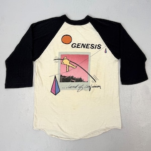 May include: Cream-colored baseball tee with black sleeves featuring a graphic from the band Genesis. The design includes a sun, a figure on a tightrope, and the words "Genesis" and "...land of confusion."