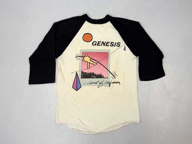 May include: Cream-colored baseball tee with black sleeves featuring a graphic from the band Genesis. The design includes a sun, a figure on a tightrope, and the words "Genesis" and "...land of confusion."
