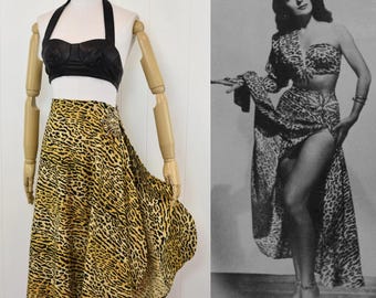 May include: A black halter-style crop top paired with a high-waisted, yellow and black leopard print skirt featuring a side embellishment. The set is displayed alongside a black and white photograph of a person wearing a similar outfit.