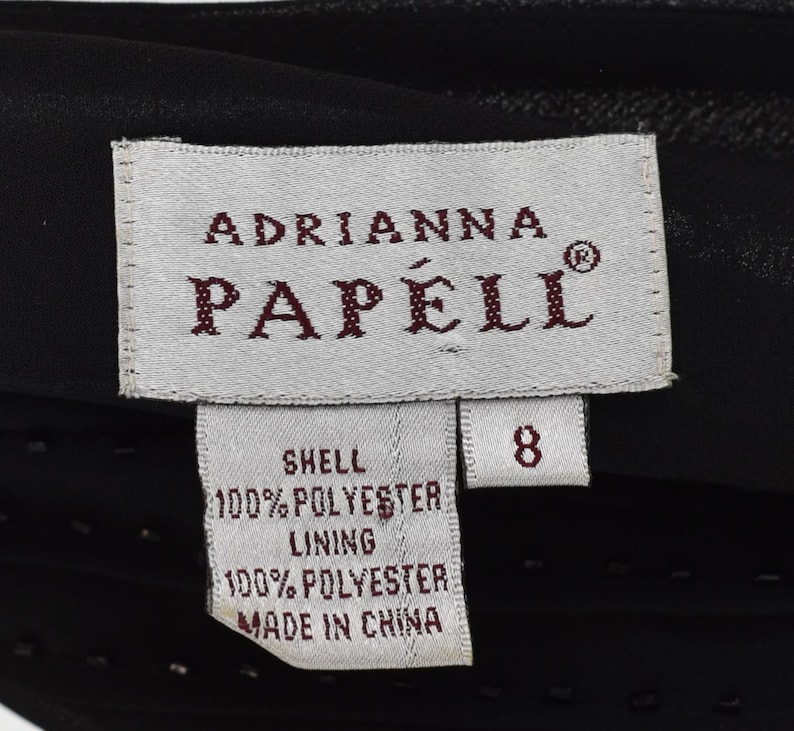 May include: A close-up of a black garment label featuring the brand name "Adrianna Papell". The label indicates the garment is size 8 and made of 100% polyester shell and lining. The label also states "Made in China."