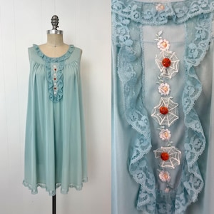 May include: A vintage, sleeveless, light blue nightgown with a round neckline and a ruffled bodice. The nightgown features lace trim along the neckline, bodice, and hem. The bodice is adorned with embroidered floral and spiderweb-like designs, and small, decorative buttons.