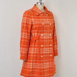 May include: An orange plaid coat with white accents, displayed on a mannequin. The coat features a double-breasted design with white buttons, a collared neckline, and a matching belt. The coat's plaid pattern is a classic design.