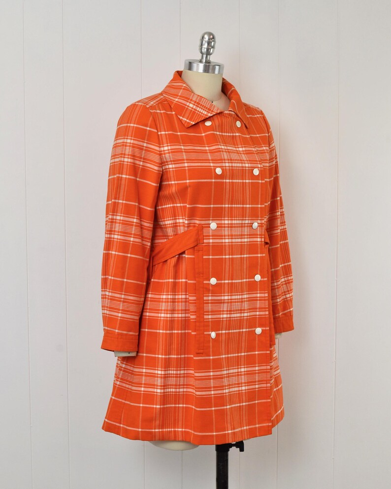 May include: An orange plaid coat with white accents, displayed on a mannequin. The coat features a double-breasted design with white buttons, a collared neckline, and a matching belt. The coat's plaid pattern is a classic design.