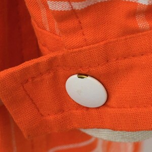 May include: Close-up of an orange fabric with white stripes and a white button. The button is oval-shaped and attached to a cuff. The fabric appears to be a shirt or blouse.