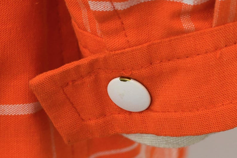 May include: Close-up of an orange fabric with white stripes and a white button. The button is oval-shaped and attached to a cuff. The fabric appears to be a shirt or blouse.