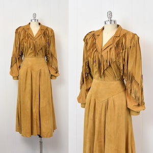 May include: A tan suede two-piece outfit on a mannequin. The top has long sleeves and fringe across the shoulders and chest. The skirt is a midi length with a high waist and pleated design.