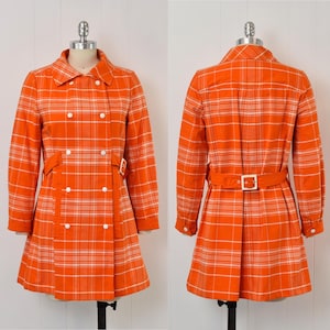 May include: An orange plaid coat with a double-breasted design, white buttons, and a matching belt. The coat features long sleeves and a classic collar. The coat is displayed on a mannequin.