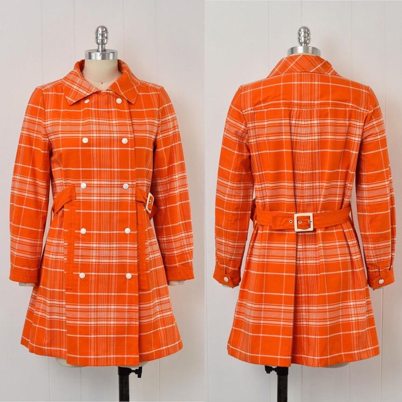 May include: An orange plaid coat with a double-breasted design, white buttons, and a matching belt. The coat features long sleeves and a classic collar. The coat is displayed on a mannequin.