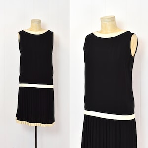 May include: A black sleeveless dress with a white trim around the neckline and hem. The skirt is pleated and falls to the knee.