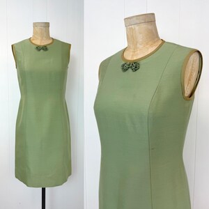 May include: A sleeveless, light green dress displayed on a mannequin. The dress features a round neckline and a decorative embellishment at the chest. The edges of the dress are trimmed with a darker green color. The dress is a vintage style.