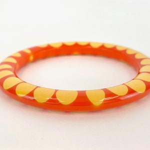 May include: Orange bangle bracelet with a repeating pattern of yellow petal-like shapes. The bracelet is a solid, circular band, likely made of resin or a similar material. The design is simple and eye-catching.