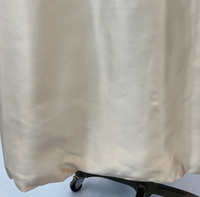 Op de afbeelding: Een close-up van de onderkant van een cr&egrave;mekleurige satijnen jurk. De jurk hangt aan een zwarte metalen standaard met wielen.