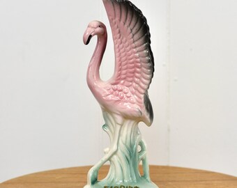 May include: Pink ceramic flamingo figurine with black wings, standing on a green base with the word "FLORIDA" written on the front.