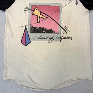May include: Cream-colored raglan t-shirt with black sleeves. The front features a graphic of a tightrope walker over a city skyline, framed within a pink and gray Polaroid-style image. Below the image, the text reads "...land of confusion."