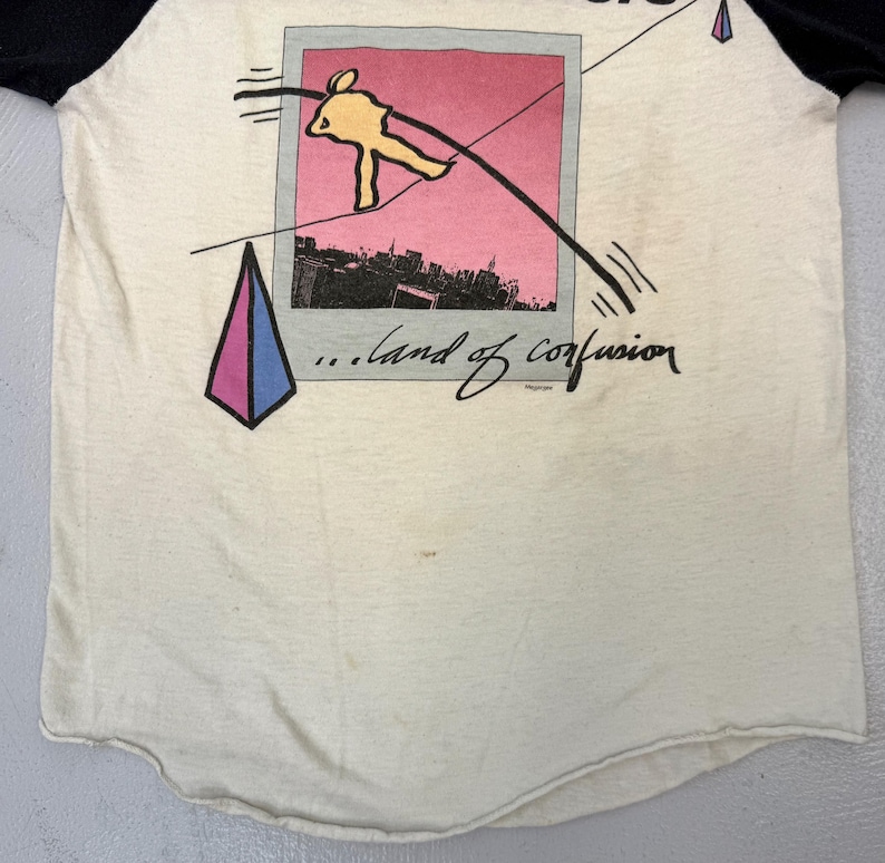 May include: Cream-colored raglan t-shirt with black sleeves. The front features a graphic of a tightrope walker over a city skyline, framed within a pink and gray Polaroid-style image. Below the image, the text reads "...land of confusion."