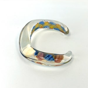 May include: A clear glass bangle bracelet with a colorful abstract design in shades of blue, yellow, and orange.