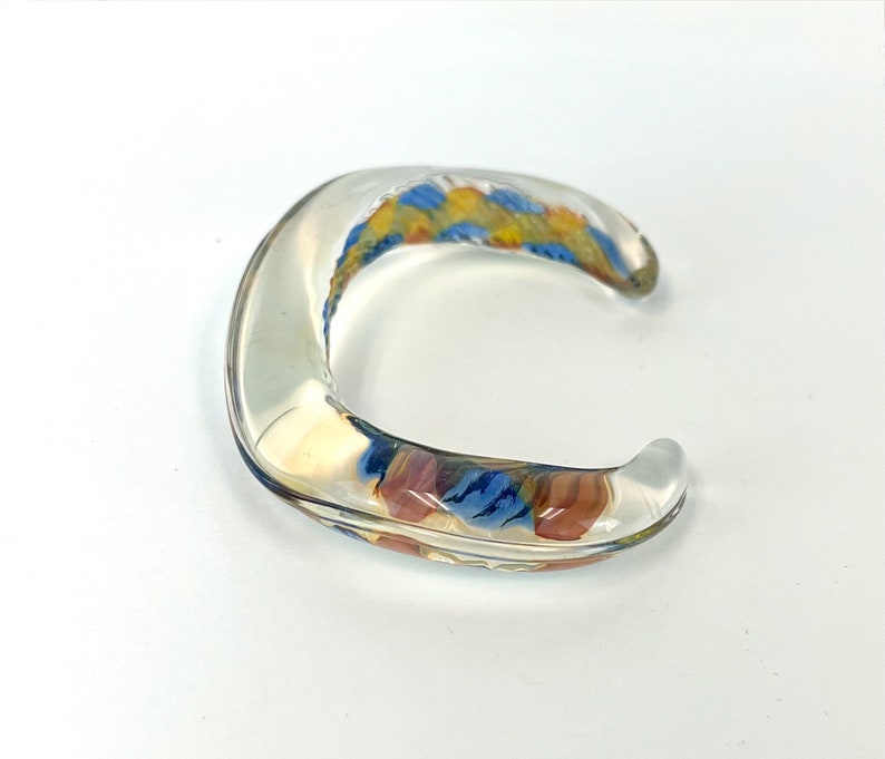 May include: A clear glass bangle bracelet with a colorful abstract design in shades of blue, yellow, and orange.