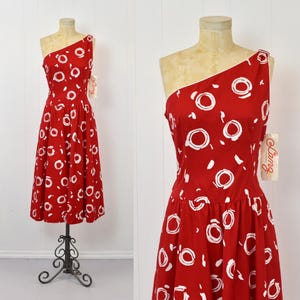 May include: A red one-shoulder dress with a white abstract print. The dress has a fitted bodice and a full skirt. The dress is made of a lightweight fabric and has a vintage feel.