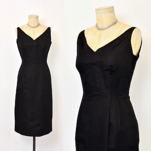 May include: A sleeveless, black dress with a V-neckline is displayed on a mannequin. The dress has a fitted bodice and a straight skirt, and is accessorized with a silver necklace. The dress is a classic, elegant style.