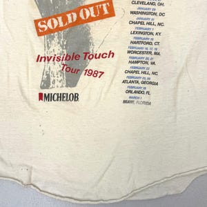May include: Cream-colored vintage t-shirt featuring the text "SOLD OUT" in an orange rectangle, with "Invisible Touch Tour 1987" in red. The shirt also lists tour dates and locations, and the Michelob logo.