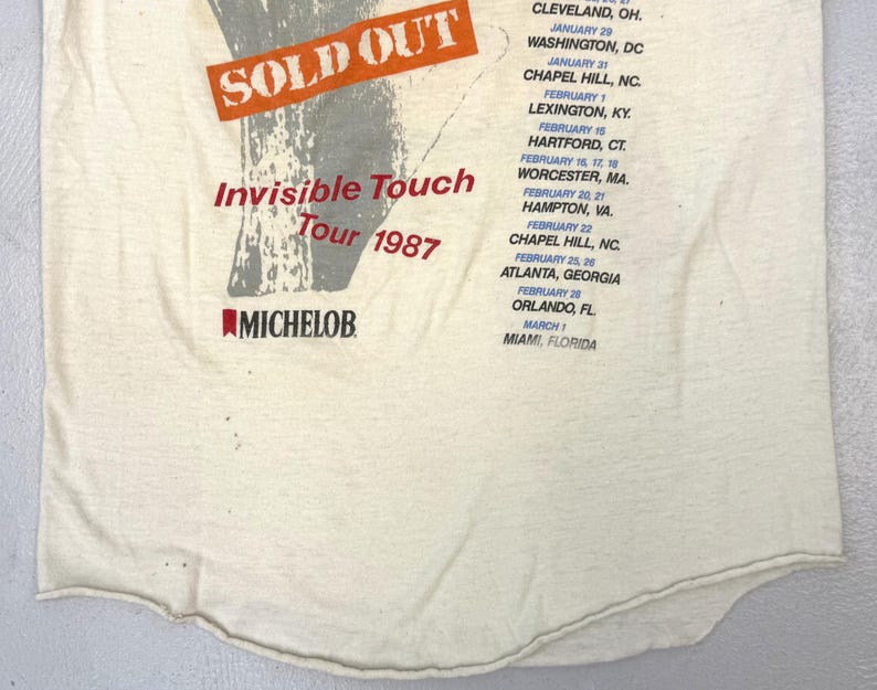 May include: Cream-colored vintage t-shirt featuring the text "SOLD OUT" in an orange rectangle, with "Invisible Touch Tour 1987" in red. The shirt also lists tour dates and locations, and the Michelob logo.