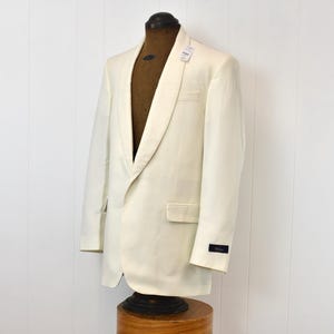 May include: Ivory-colored tuxedo jacket with a shawl collar, displayed on a mannequin. The jacket has a single-button closure and a pocket on the left side. A small tag is attached to the collar.