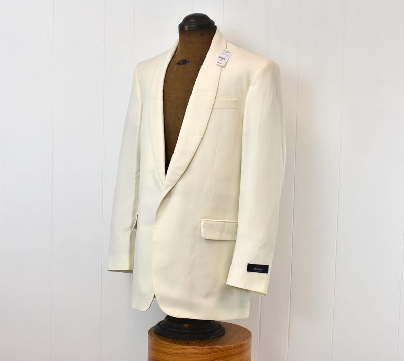 May include: Ivory-colored tuxedo jacket with a shawl collar, displayed on a mannequin. The jacket has a single-button closure and a pocket on the left side. A small tag is attached to the collar.