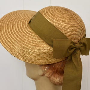 May include: A straw hat with a wide brim and a rounded crown. The hat features a wide olive green ribbon that wraps around the crown and ties into a bow at the back. The hat is displayed on a mannequin head.