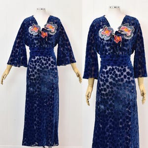 May include: A floor-length navy blue velvet dress featuring a burnout floral pattern, wide bell sleeves, and a V-neckline. The bodice is adorned with three-dimensional fabric flowers in shades of pink, orange, and blue with green accents.