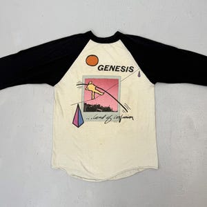 May include: Cream-colored baseball tee with black sleeves. The shirt features the band name "GENESIS" above a graphic of a pink and orange image with a flying figure and the words "...land of confusion".