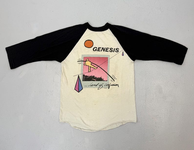 May include: Cream-colored baseball tee with black sleeves. The shirt features the band name "GENESIS" above a graphic of a pink and orange image with a flying figure and the words "...land of confusion".