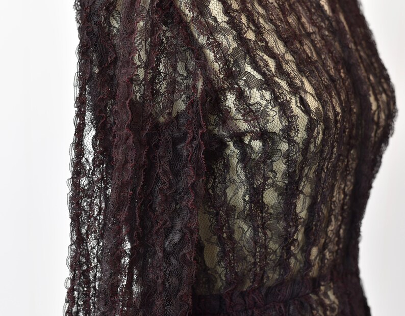 May include: A close-up of a dark burgundy lace fabric with a delicate floral pattern. The fabric is sheer and has a slightly ruffled texture.