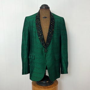 Może przedstawiać: Zielony blazer z czarnym satynowym kołnierzem ozdobionym mieniącymi się cekinami. Blazer ma pojedyncze zapięcie na guzik i kieszeń po lewej stronie.