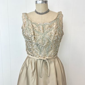 Op de afbeelding: Een mouwloze, lange, lichtbeige satijnen jurk met een getailleerd bovenstuk en een volle rok. Het bovenstuk is versierd met een delicaat bloemenpatroon in een lichtere tint groen en zilveren pailletten.