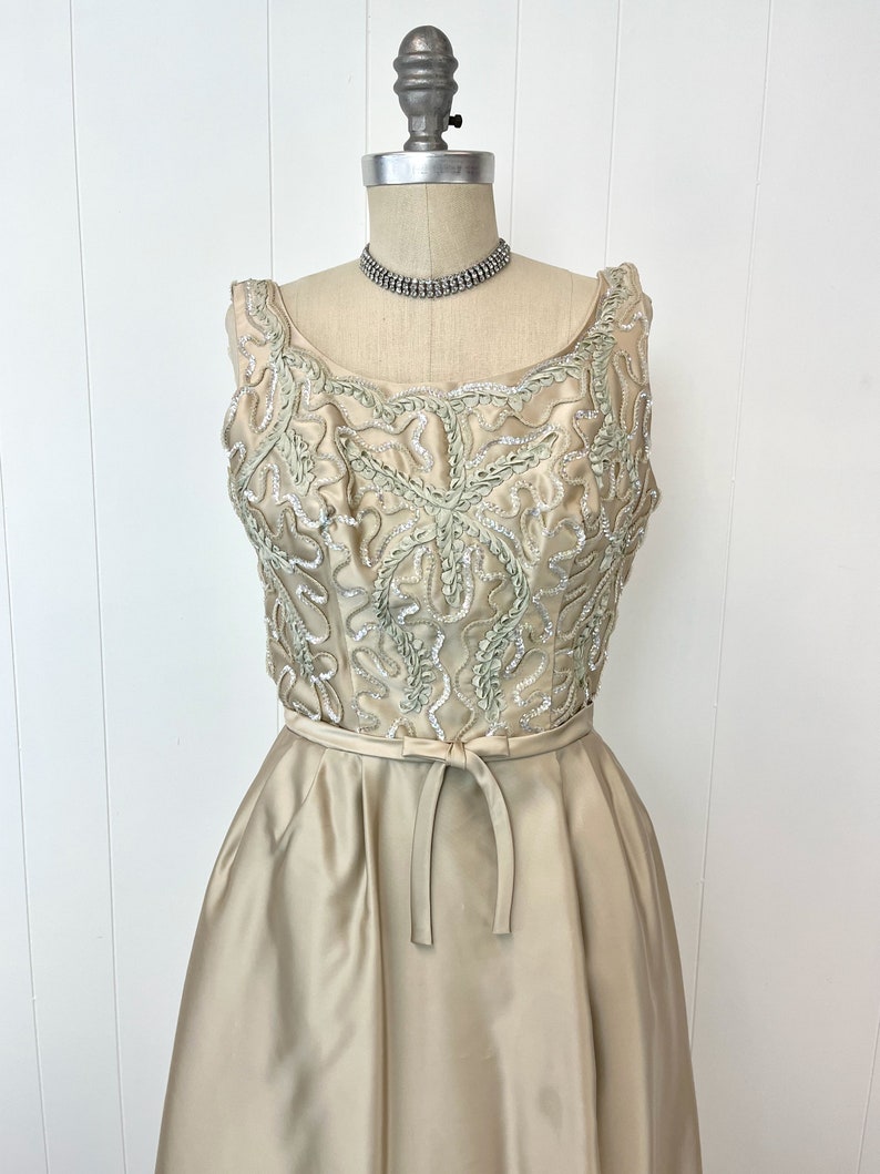Op de afbeelding: Een mouwloze, lange, lichtbeige satijnen jurk met een getailleerd bovenstuk en een volle rok. Het bovenstuk is versierd met een delicaat bloemenpatroon in een lichtere tint groen en zilveren pailletten.