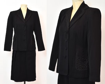 May include: A vintage black skirt suit displayed on a mannequin. The suit features a tailored blazer with decorative stitching around the collar and pockets, paired with a matching knee-length skirt. The blazer has button closures. The suit is made of a solid black fabric.