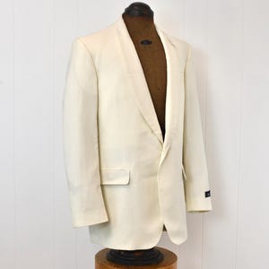 May include: An off-white tuxedo jacket with a shawl collar, displayed on a vintage mannequin. The jacket has a single-button closure and a pocket on the left side. The jacket is made of a smooth fabric.