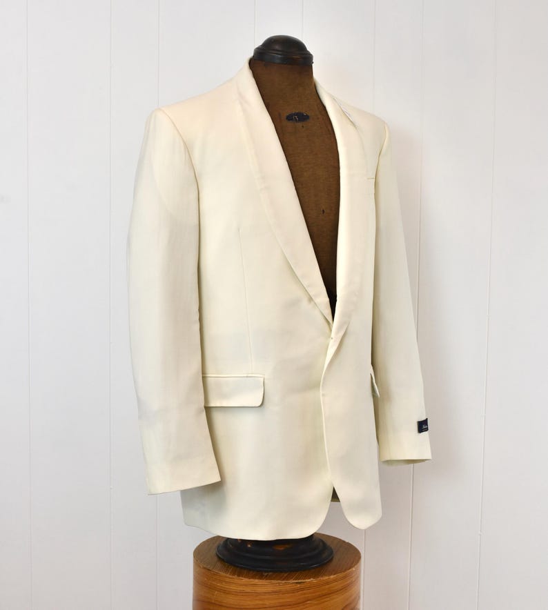 May include: An off-white tuxedo jacket with a shawl collar, displayed on a vintage mannequin. The jacket has a single-button closure and a pocket on the left side. The jacket is made of a smooth fabric.