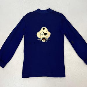 May include: A long-sleeved, navy blue shirt with a crew neck. The shirt features an embroidered pirate face on the back, with a white hat, eye patch, and mustache. The sleeves have ribbed cuffs.