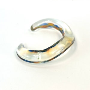 May include: A clear glass bangle with a wavy, organic shape. The bangle has a subtle iridescent sheen with hints of blue and orange.