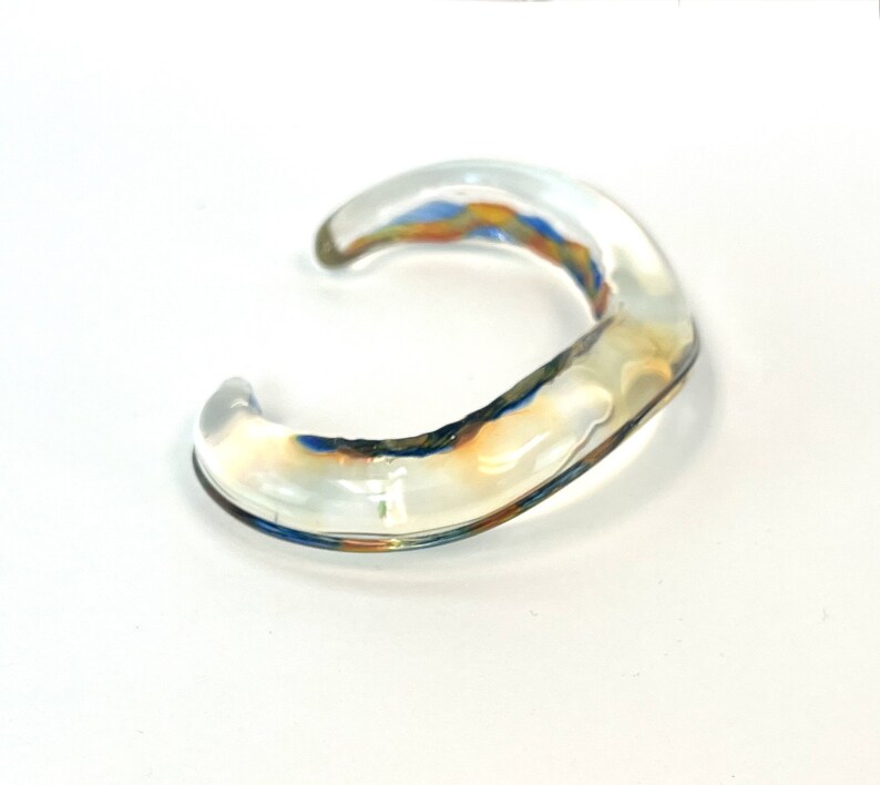 May include: A clear glass bangle with a wavy, organic shape. The bangle has a subtle iridescent sheen with hints of blue and orange.